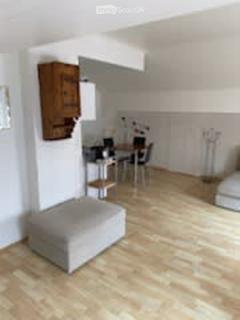 A modern, well-lit room with light wooden flooring, a grey sofa, a small dining table with chairs, a wooden cabinet, and a wall-mounted lamp.