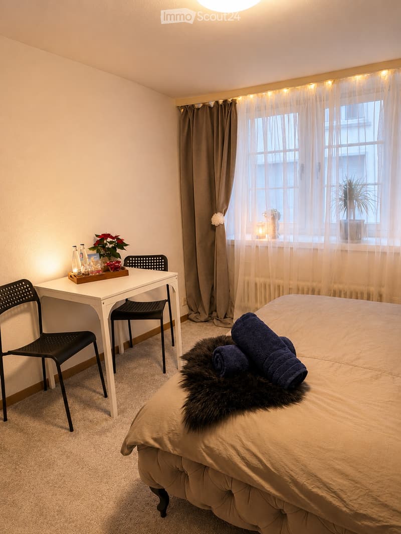 Cozy bedroom with a bed, a small table with chairs, and a window with curtains. The bed has a fur throw and blue towels, and there is a small plant on the windowsill.