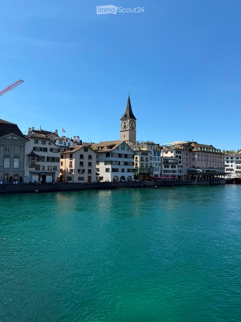 A view of a cityscape with a prominent clock tower overlooking a calm river, surrounded by multi-story buildings with various architectural styles.