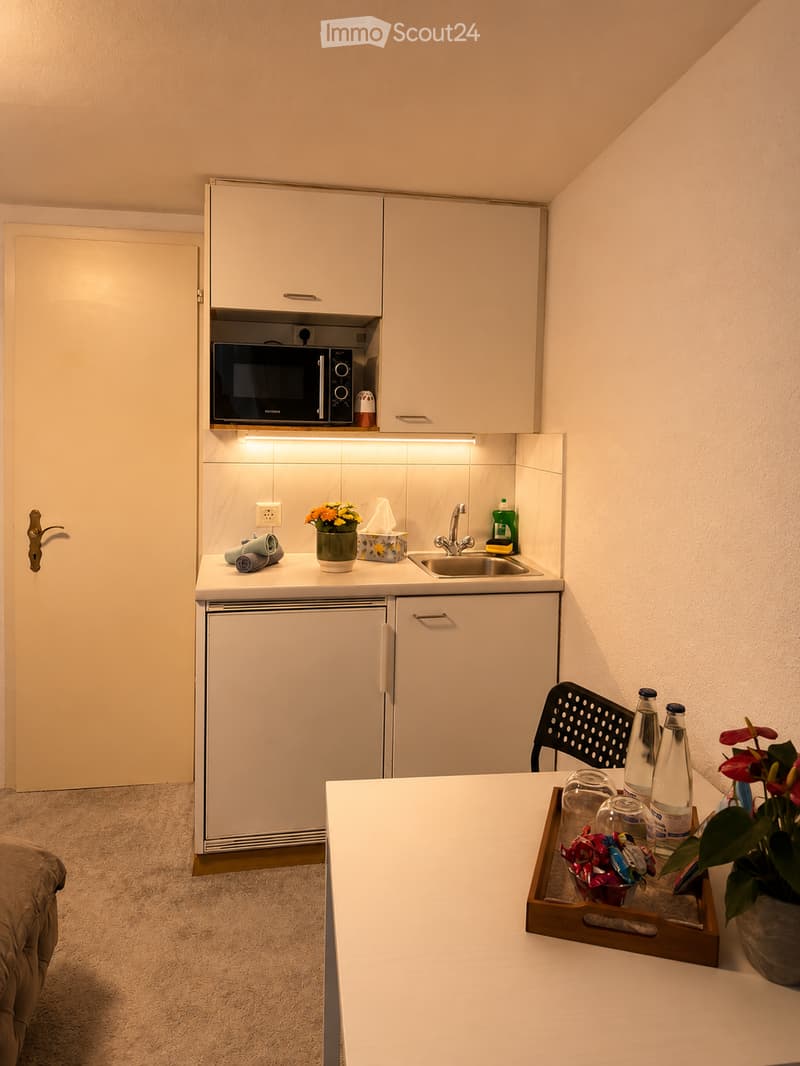 Compact, modern kitchen area with white cabinets, microwave, sink, and small refrigerator. A small table with a tray of drinks and snacks, a chair, and decorative flower pots are present.
