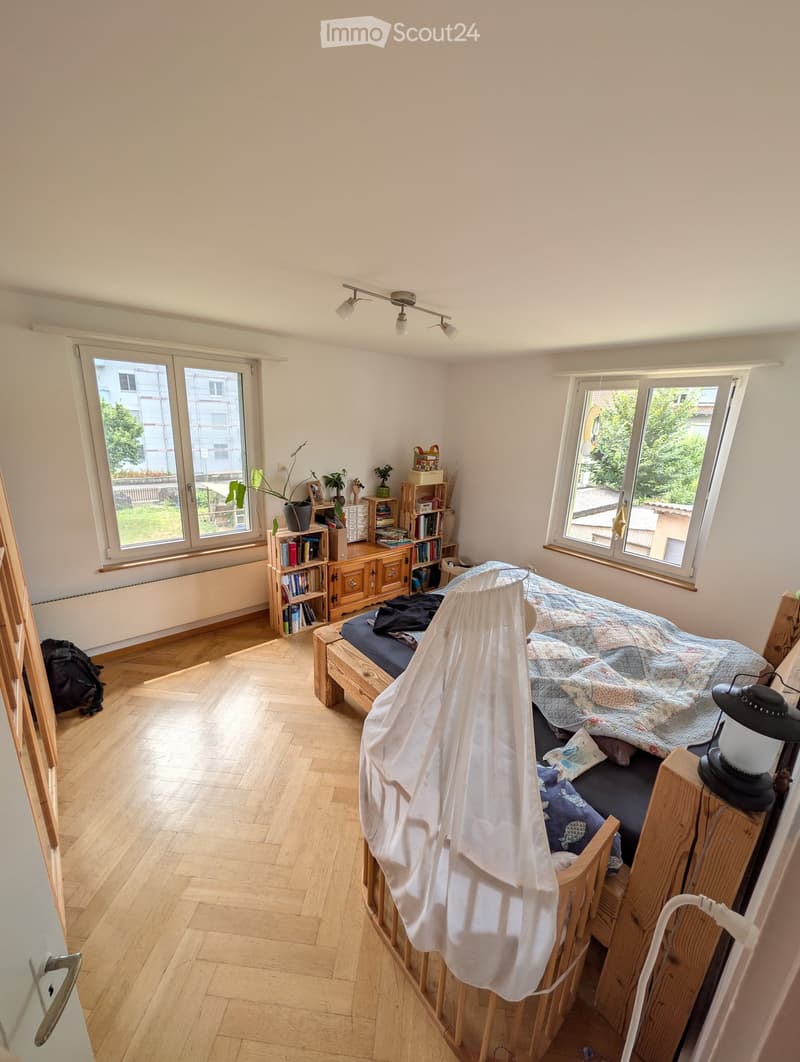 Bright bedroom with wooden flooring, large windows, a bed with a patterned quilt, bedside table with a lamp, bookshelf, and potted plants.