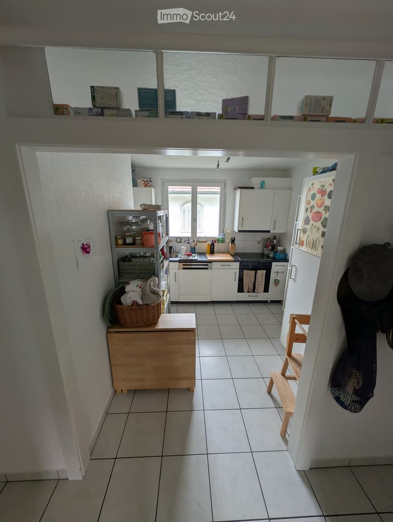 Bright kitchen with white cabinets, tiled floor, and various appliances including a refrigerator, oven, and dishwasher. A wooden stool and basket are placed near the entrance.