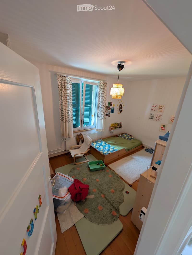 Cozy children's bedroom with a single bed, a small chair, a green rug, and a storage unit. The room features wooden flooring, a window with curtains, and various toys and decorations.