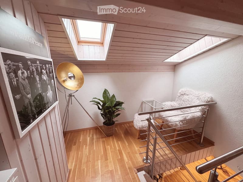 A modern hallway with wooden flooring, white walls, and skylights. Features include a large framed photo, a floor lamp, a potted plant, and a white fur chair on a small platform.