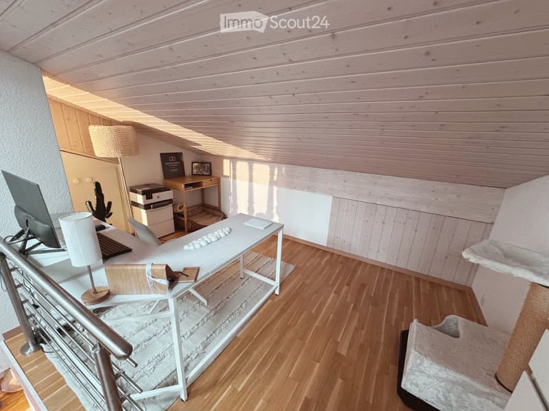 A modern, well-lit room with wooden flooring, a desk setup, a small table, and a cozy seating area with a white chair and a cushioned seat.