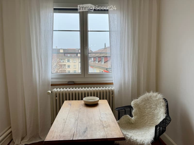 A small, unfurnished room with a wooden table, a white bowl, and a white fur-covered chair near a window with white curtains. The room has a radiator and a view of rooftops.