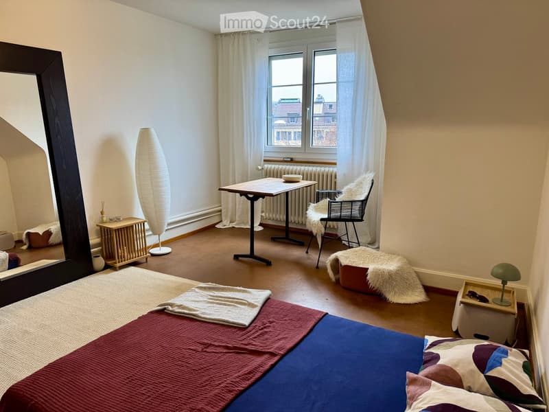 Cozy bedroom with a bed, a small table, a chair, a floor lamp, and a mirror. The room features large windows with curtains and a radiator below them.