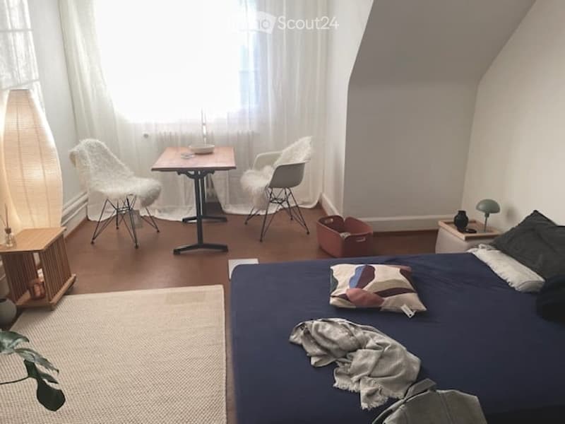 Cozy bedroom with a bed, two chairs, a small table, a floor lamp, and a plant. The room features wooden flooring and large windows with sheer curtains.