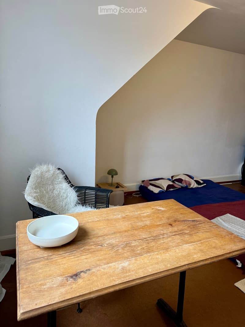 A small, cozy bedroom with a wooden table, a white bowl, a wicker chair with a fur throw, a bed with colorful pillows, and a small table lamp.
