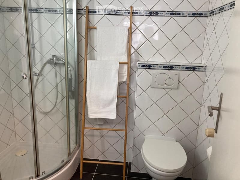 Modern bathroom with white tiled walls, a glass-enclosed shower, a toilet, and a wooden towel rack holding white towels.