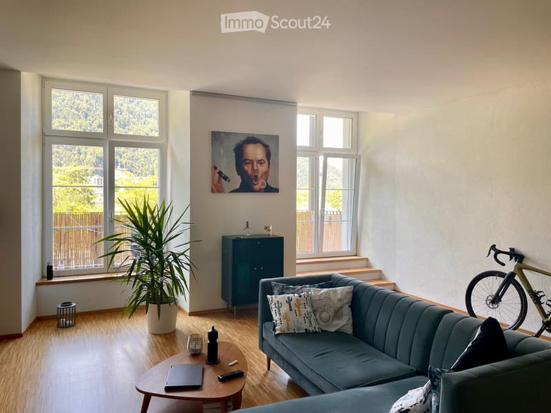 Modern living room with a grey sofa, wooden coffee table, laptop, remote control, decorative cushions, a potted plant, a cabinet, and a bicycle leaning against the wall. Large windows offer a view of greenery outside.