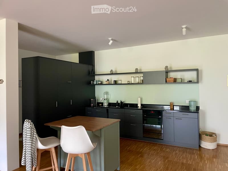 Modern kitchen with dark grey cabinets, a central island, and white stools. Appliances include a blender, oven, and sink. Shelves hold various kitchen items.
