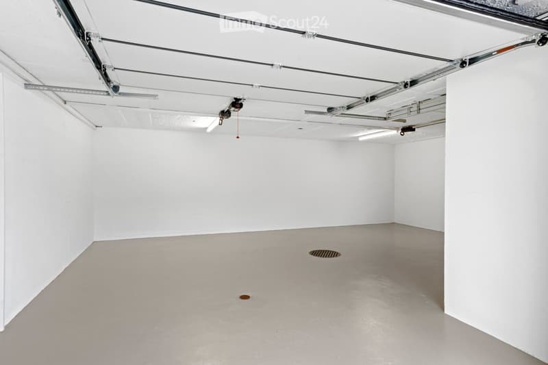 Empty room with white walls, grey concrete floor, and exposed ceiling with lighting fixtures and mechanical elements.