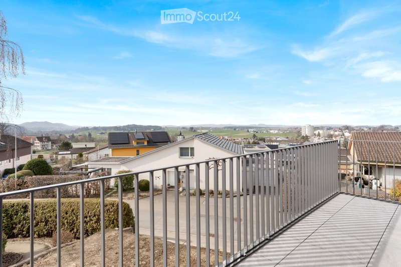 Balcony with metal railing overlooking a suburban neighborhood with houses, greenery, and distant hills under a clear blue sky.