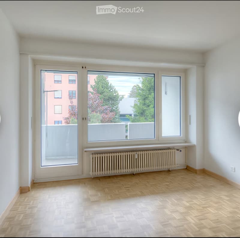 Empty room with wooden flooring, white walls, large windows, radiator, and a sliding door leading to a balcony.