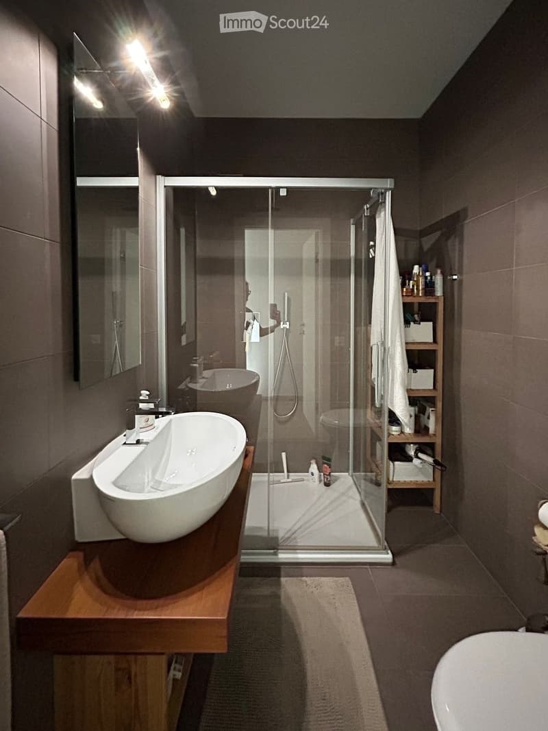 Modern bathroom with a large bathtub, sink, glass-enclosed shower, and wooden shelf holding towels and toiletries.