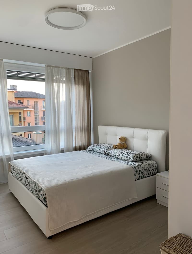 Modern bedroom with a large bed, patterned bedding, a nightstand, and a teddy bear on the bed. The room features wooden flooring, large windows with curtains, and a round ceiling light.