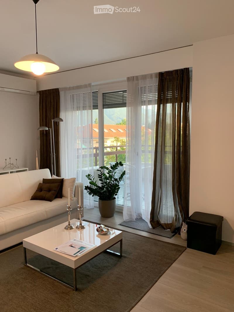 Modern living room with white sofa, coffee table, candles, and a potted plant near large windows with sheer curtains.