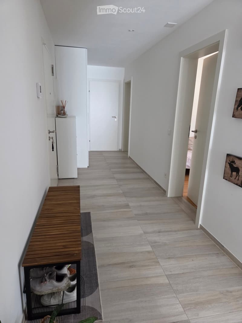 Modern hallway with light wooden flooring, white walls, and several doors leading to different rooms. A wooden bench with shoes underneath is positioned on the left side, and a small cabinet with decorative items is visible near the entrance.