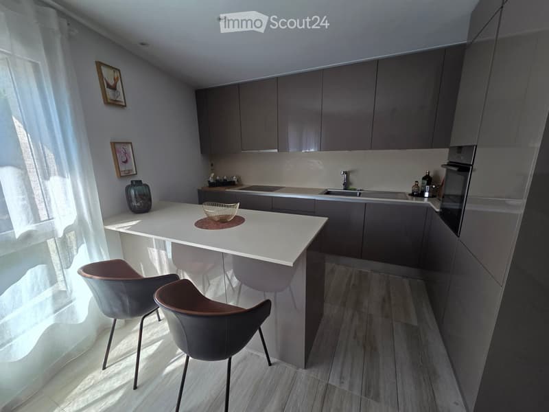 Modern kitchen with grey cabinets, white island, two chairs, sink, oven, and decorative items.