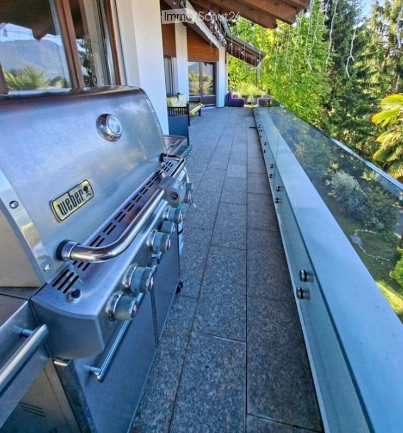 Modern balcony with a glass railing, tiled floor, and a Weber grill on the left side. The background shows a lush green view of trees and part of a house with outdoor seating.