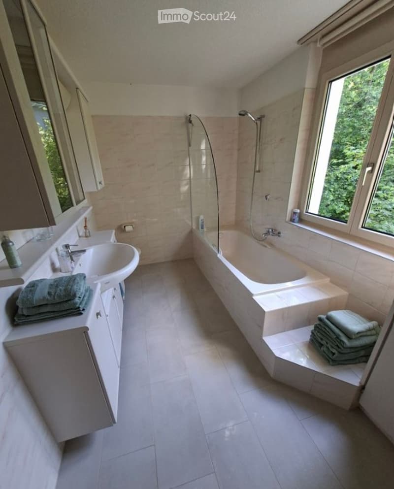 Modern bathroom with a white tiled floor, a large bathtub, a sink with a mirror, and neatly folded towels on the cabinet and bathtub steps.