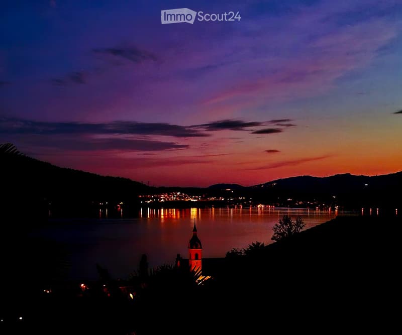 Evening view of a lake with city lights reflecting on the water, surrounded by hills and a small illuminated tower.