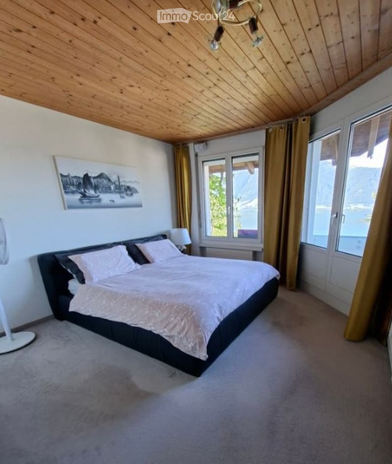 Modern bedroom with a large bed, nightstand, lamp, fan, and a scenic view through large windows with curtains.