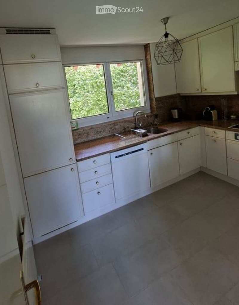 Bright kitchen with white cabinets, a central island, and a large window overlooking greenery. Features include a sink, dishwasher, and modern lighting.