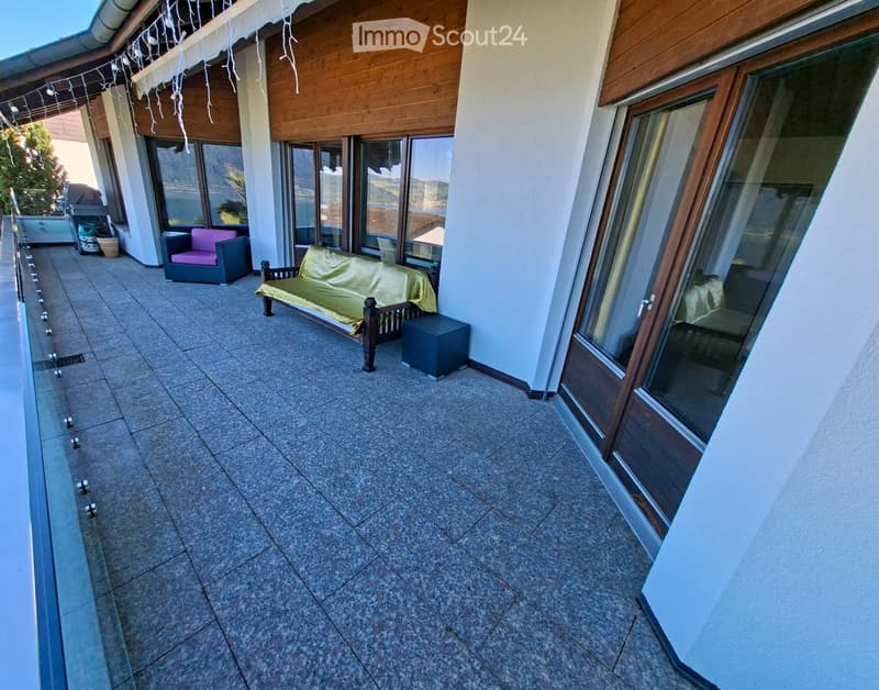 Balcony with tiled floor, large windows, sofa, chair, and bench, overlooking a scenic view.