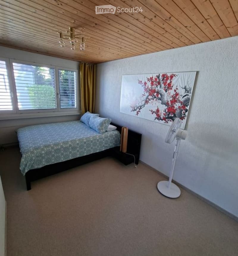 Cozy bedroom with a wooden ceiling, a bed with patterned bedding, a nightstand, a fan, and a large abstract painting on the wall.