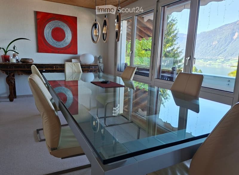 Modern dining room with a large glass table, leather chairs, wooden sideboard, potted plant, and decorative artwork. Large windows offer a scenic mountain view.