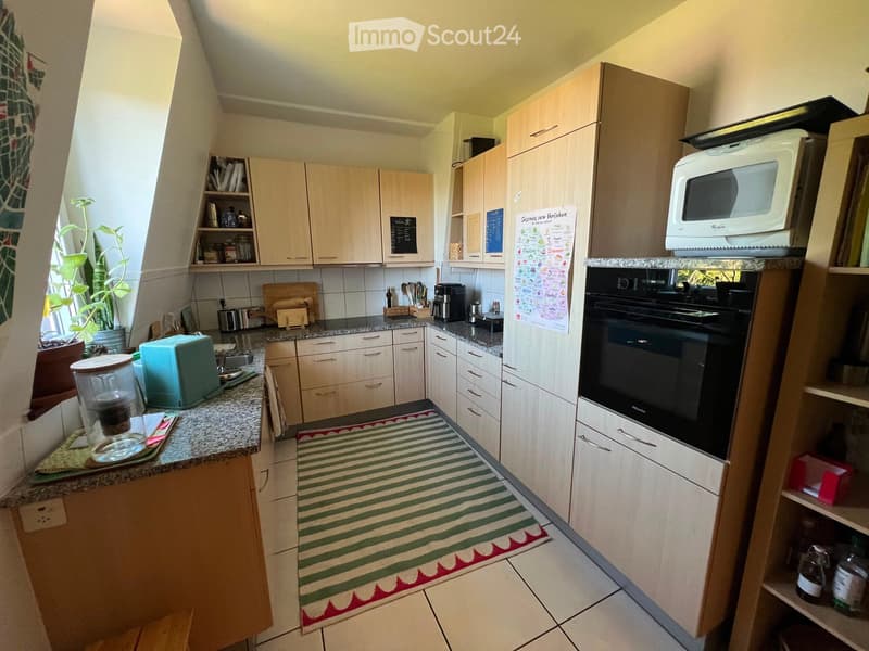 Bright kitchen with light wood cabinets, granite countertops, and various appliances including an oven, microwave, refrigerator, coffee maker, and toaster. A green and red striped rug is on the floor, and a potted plant is near the window.