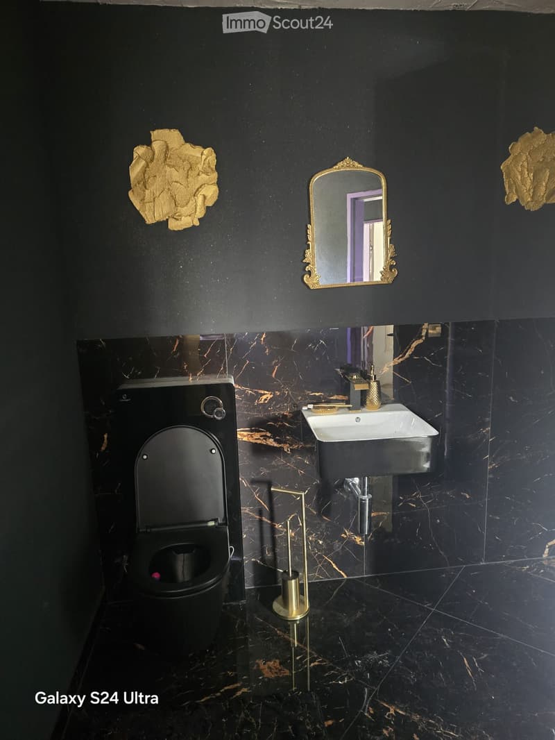 Modern bathroom with black marble walls and floor, featuring a sleek black toilet, a white sink with gold fixtures, and an ornate gold-framed mirror.