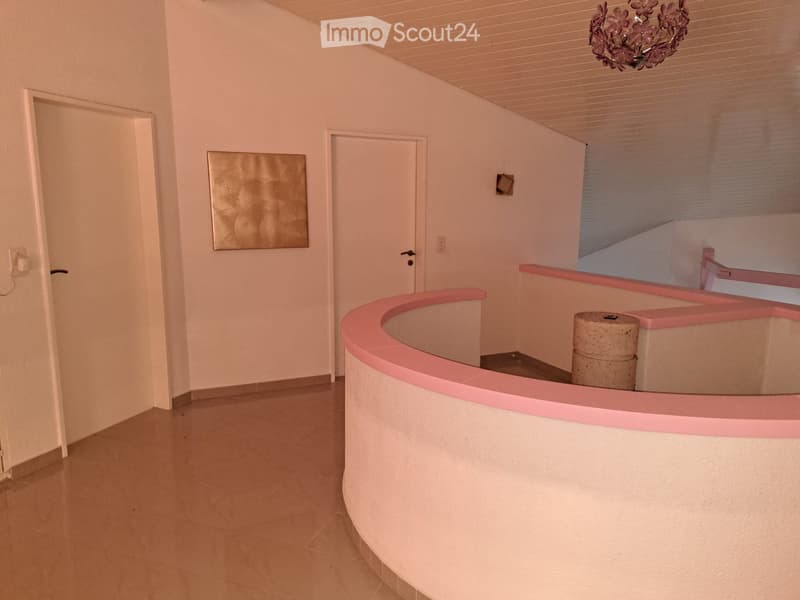 Empty hallway with light-colored walls, tiled floor, and a curved pink railing leading to a staircase. There are two doors and a decorative wall piece visible.