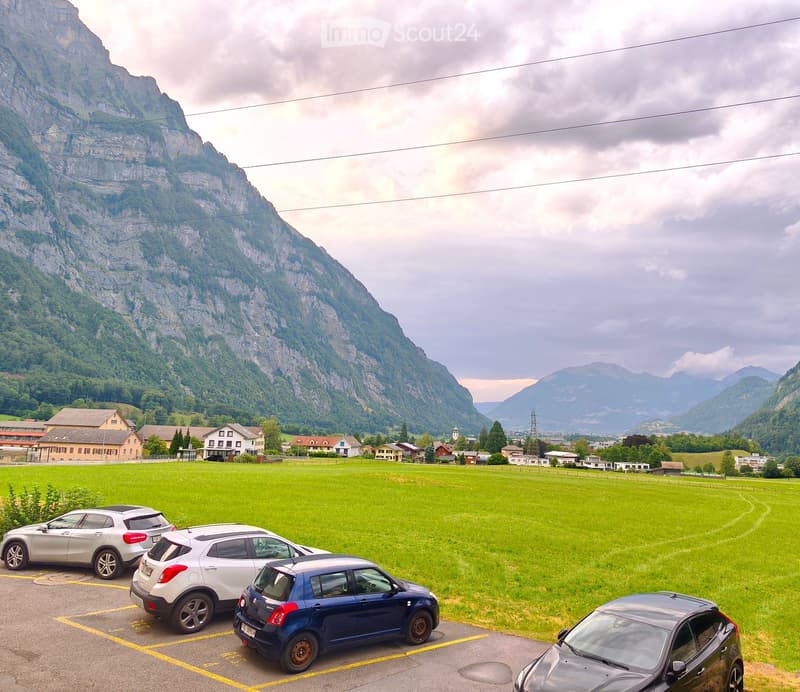 Parking lot with several cars, surrounded by a green field and distant mountains.