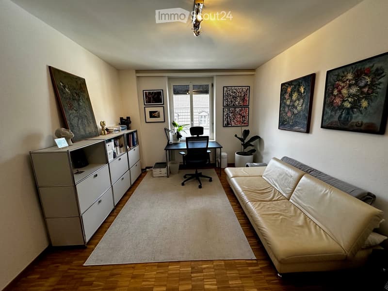 A modern living room with a cream-colored sofa, a black office chair, a desk, a cabinet, and several picture frames on the walls.