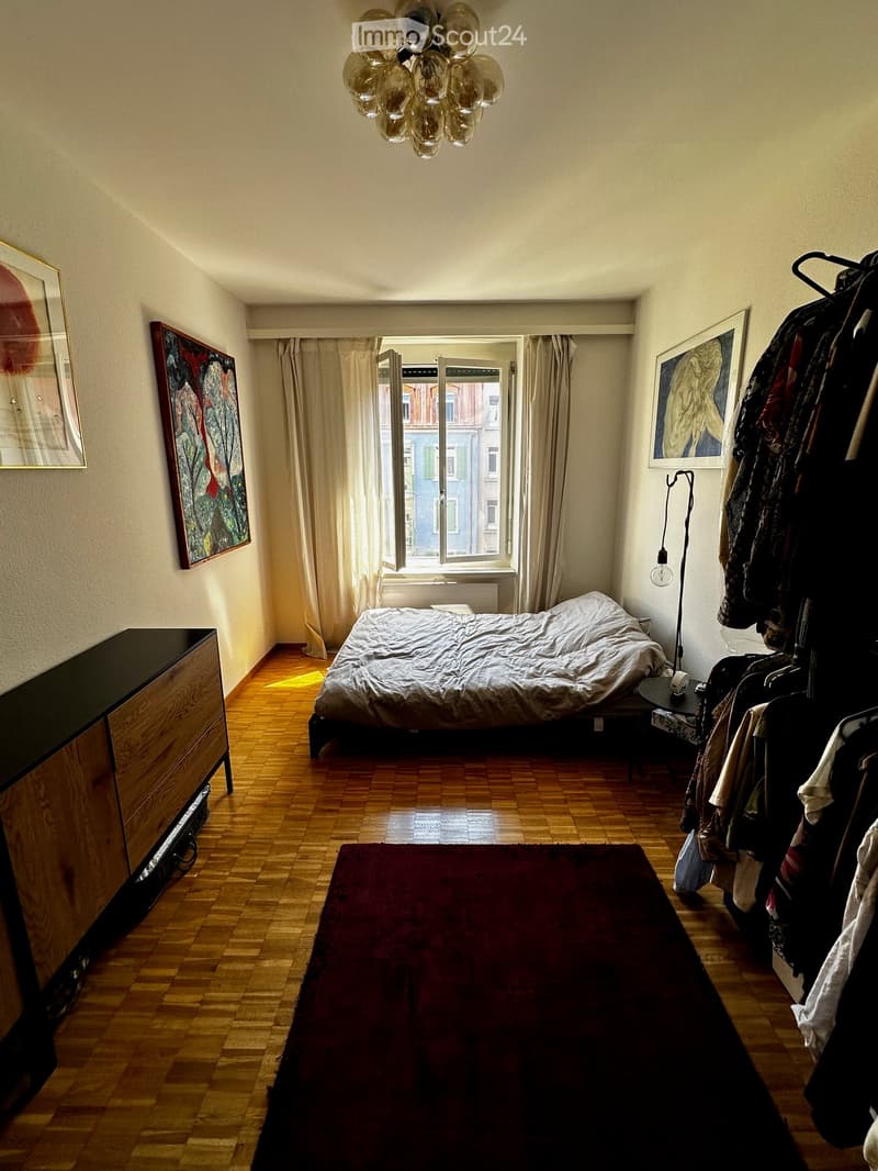 Cozy bedroom with wooden flooring, a large bed, a nightstand, a wardrobe, and artwork on the walls. The room features a large window with curtains and a red rug on the floor.