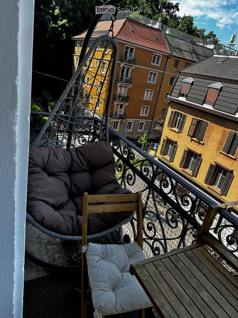 Balcony with a wooden table, a cushioned chair, and a hanging wicker chair, overlooking a cityscape with yellow buildings and balconies.