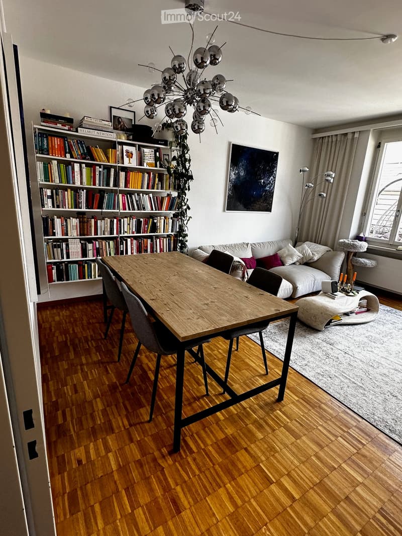Modern living room with wooden flooring, a large bookshelf filled with books, a wooden dining table with chairs, a grey sofa with cushions, a floor lamp, and a cat tree near the window.