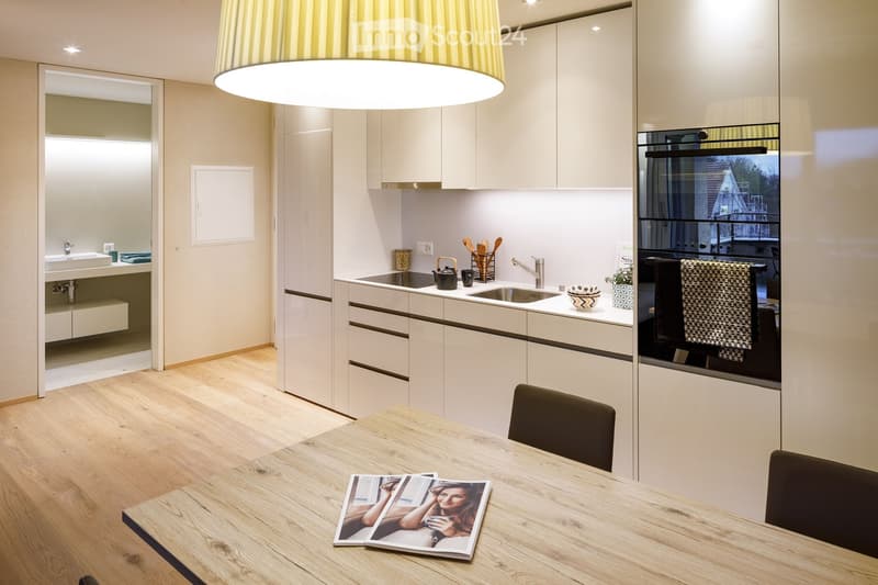 Modern kitchen with white cabinets, wooden table, and various appliances including a sink, oven, and faucet. A magazine is placed on the table.