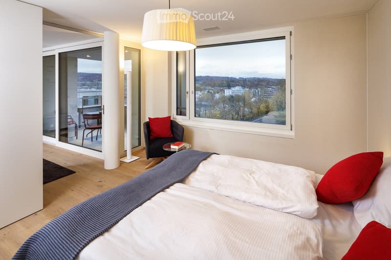 Modern bedroom with a large window offering a scenic view, a neatly made bed with red pillows, a small seating area with a chair and table, and a hanging lamp.