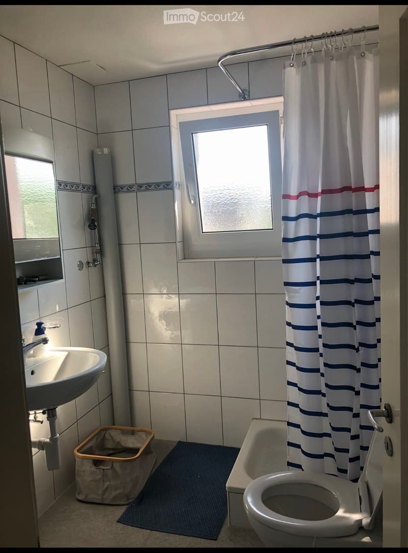 Bathroom with a sink, toilet, bathtub, and shower curtain. Features tiled walls, a window, and a small trash bin.