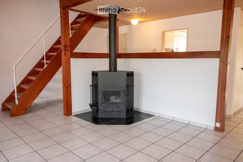 A hallway with a tiled floor, a wooden staircase leading upwards, and a black wood-burning stove with a chimney. The walls are white, and there is a light fixture on the ceiling.