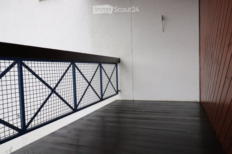 Balcony with dark wooden decking, metal railing with geometric pattern, and white textured wall in the background.