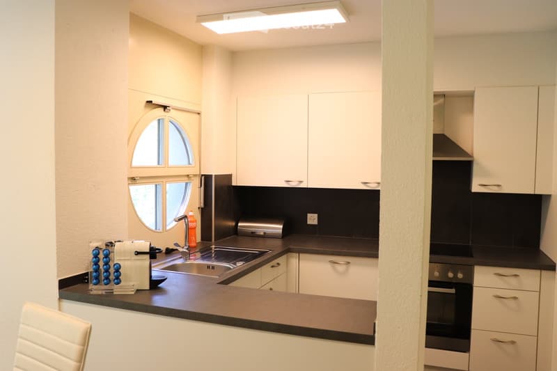 Modern kitchen with white cabinets, black countertops, stainless steel sink, and appliances including a coffee maker and toaster. Features a round window above the sink and a sleek design.