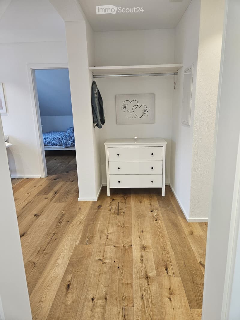 A small, neatly arranged room with light wooden flooring, white walls, a chest of drawers, and a picture frame on the wall. A closet with a hanging garment is visible, and an open doorway leads to another room with a bed.