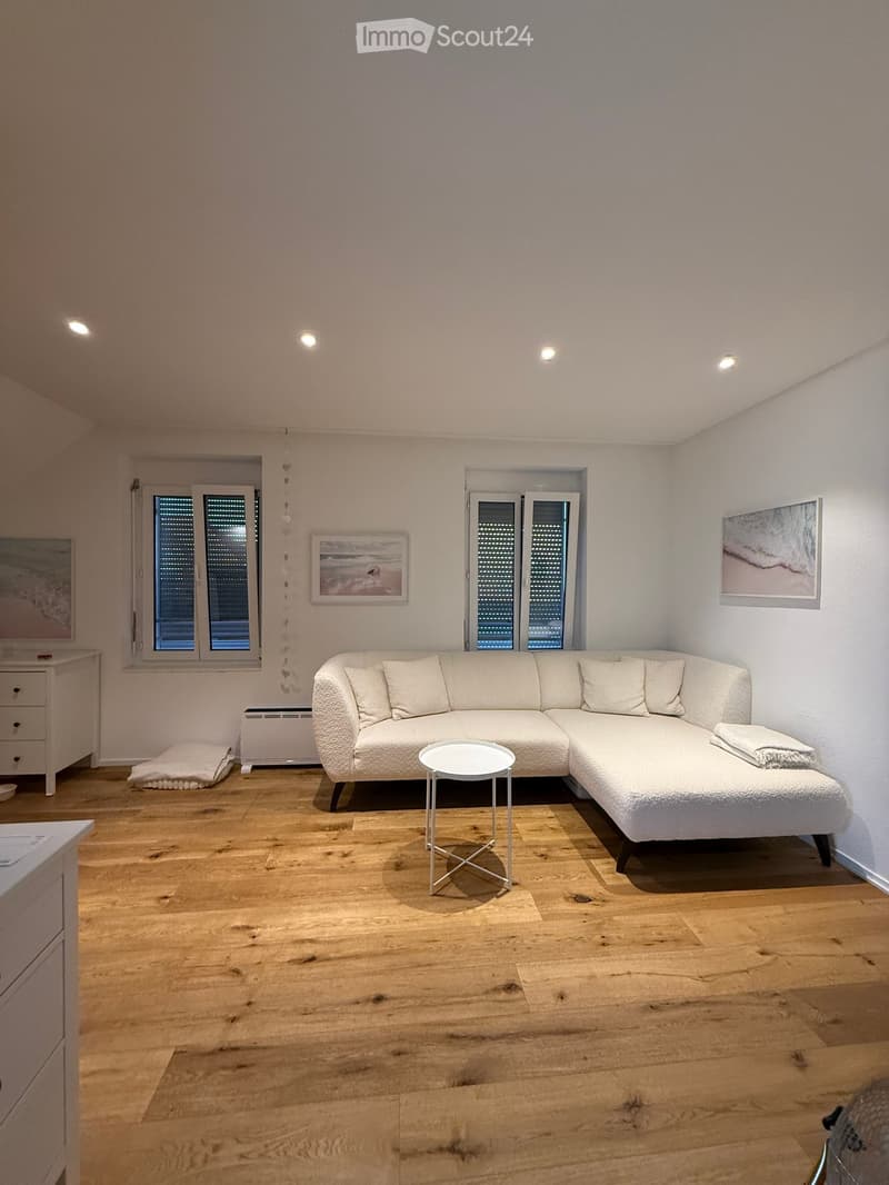 Modern living room with light wooden flooring, white walls, and large windows. Features a white sectional sofa, a small round coffee table, a side table, and picture frames on the walls.