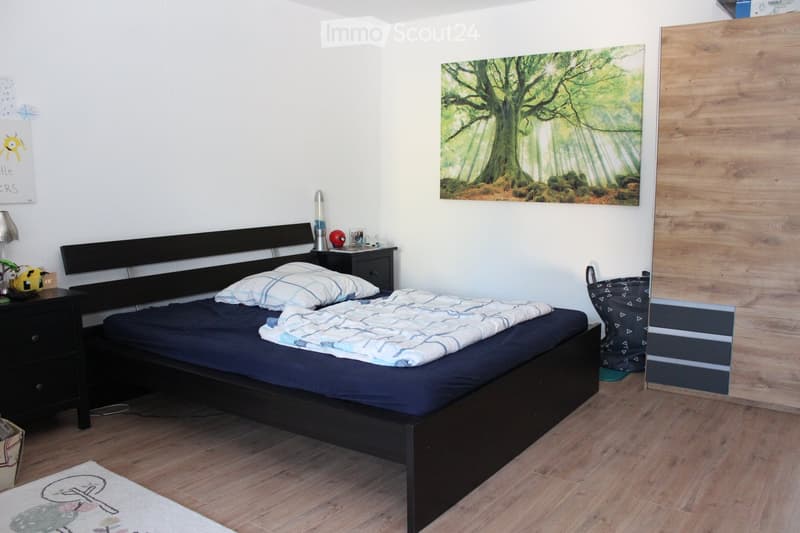 A modern bedroom with a dark wooden bed, nightstand, lamp, and a large forest-themed picture on the wall. The room features light wooden flooring and a small rug near the bed.