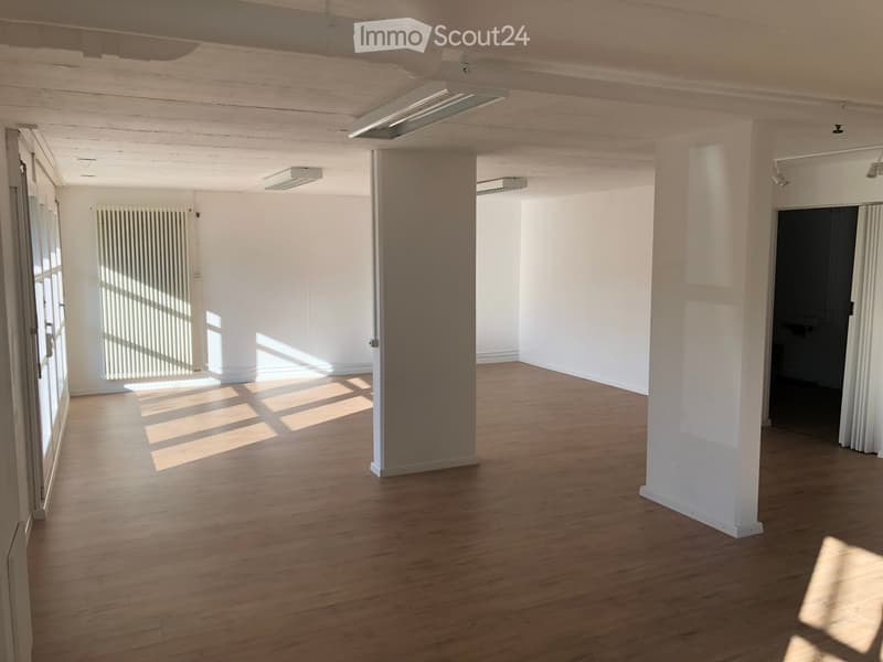 Empty room with wooden flooring, white walls, and large windows allowing natural light. Features include a radiator, a door, and exposed ceiling beams.